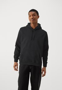 Black hoodie with a front pocket, drawstring hood, and ribbed cuffs. Worn with dark denim pants, featuring a smooth, soft texture.