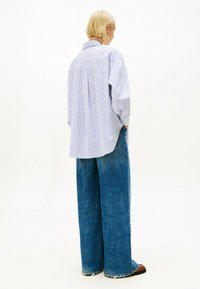 Een blonde persoon staat met zijn rug naar de camera en draagt een losse blauw gestreepte blouse, een wijde spijkerbroek en bruine schoenen tegen een witte achtergrond.
