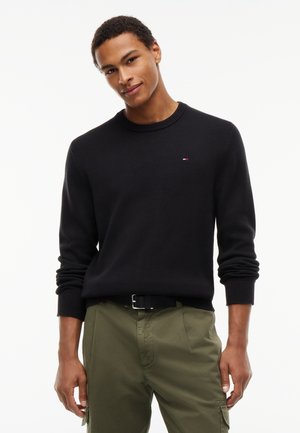 Mladý muž oblečený v čiernom pulóvri s dlhým rukávom a olivovo zelených nohaviciach stojaci na bielom pozadí.