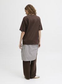 Bruine oversized t-shirt, grijze geruite rok en bruine wijdvallende broek. Kenmerken zijn zachte stof en een relaxte pasvorm. Krullend haar zichtbaar.