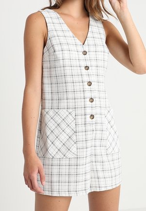 Sleeveless white dress with black plaid pattern, front buttons, and two large front pockets, worn by a standing person.