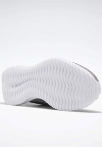 Semelle blanche texturée d'une chaussure de sport grise inclinée sur une surface blanche, mettant en valeur le motif de la bande de roulement pour l'adhérence et le soutien.