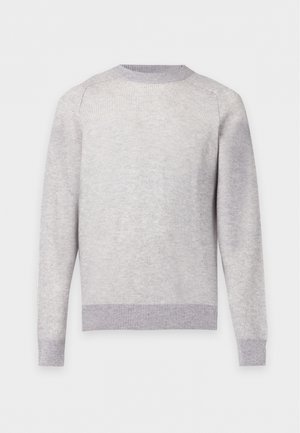 Homme portant un pull gris clair, un pantalon sombre et des mocassins marron et beige, debout avec une main dans la poche sur fond uni.