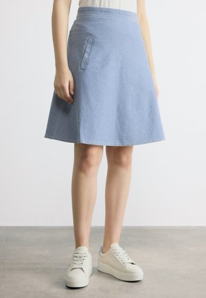 Femme portant une jupe trapèze rayée bleu et blanc longueur genou avec poches latérales et des baskets blanches, debout sur un sol gris.