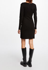 Černé šaty s dlouhým rukávem a přiléhavým živůtkem, mírně rozšířenou sukní, doplněné černými koženými botami do kolen. Hladká textura, minimalistický design.