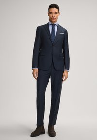 Abito navy con vestibilità sartoriale, chiusura a due bottoni, rever a punta e tasche laterali. Abbinato a una camicia azzurro chiaro e stivali marrone scuro.