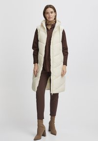 Gilet rembourré en crème clair avec une capuche, présentant un motif matelassé et une fermeture éclair sur le devant. Porté par-dessus une chemise marron et un pantalon marron assorti.