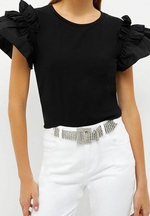 Mujer vestida con una blusa negra de manga corta con volantes y pantalones blancos con un cinturón ancho de pedrería con hebilla grande.