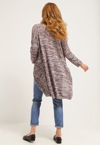 Donna con capelli rossi mossi, che indossa un lungo cardigan viola melange, jeans blu arrotolati e ballerine nere, in piedi davanti a uno sfondo semplice.