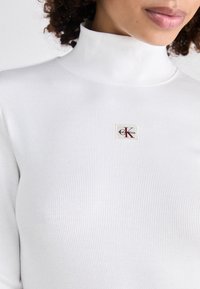 Witte geribde turtleneck met een getailleerd ontwerp, voorzien van een klein label met een rood "K"-logo op een crèmekleurige achtergrond. Zacht, gestructureerd materiaal.