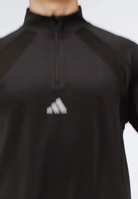 Zwarte sporttop met lange mouwen en een kwart ritssluiting, met het witte Adidas-logo op de borst, gedragen door een onzichtbaar persoon.
