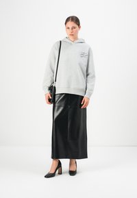 Donna che indossa un hoodie grigio chiaro, una gonna midi in pelle nera, tacchi neri e tiene una piccola borsa a tracolla nera, in piedi su uno sfondo bianco.