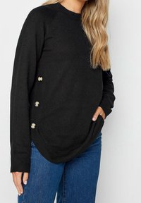 Svart stickad tröja med rund halsringning, raglanärmar och dekorativa knappar på sidan, med en krökt fåll och avslappnad passform.