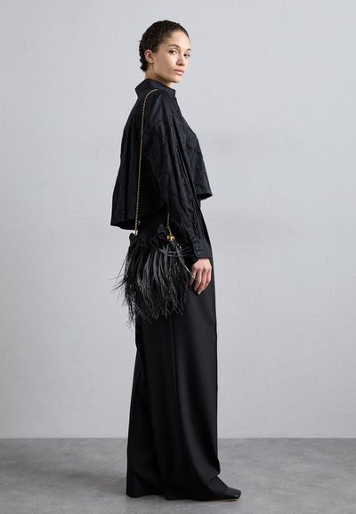 Cropped top nero con motivo a diamante, abbinato a una lunga gonna nera fluida. Tiene in mano una borsa nera con frange e tracolla dorata.