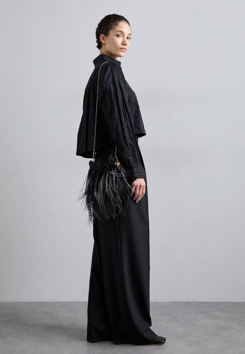 Blouse noire courte avec motif en losange, associée à une longue jupe noire fluide. Tenant un sac à franges noir avec une bandoulière en chaîne dorée.