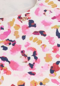 Tissu à motif floral en rose, jaune et bleu sur un fond crème. Présente un petit accent de logo doré près du col.