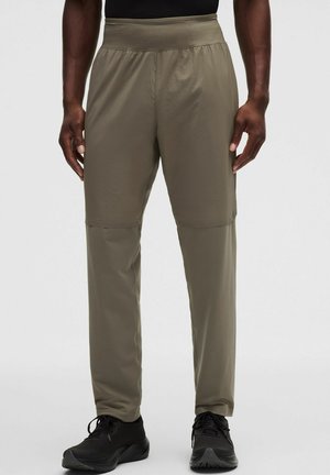 Mann trägt olivgrüne Convertible-Hose mit schwarzen Turnschuhen und schwarzem Oberteil, gezeigt von der Taille bis zu den Füßen vor einfarbigem Hintergrund.