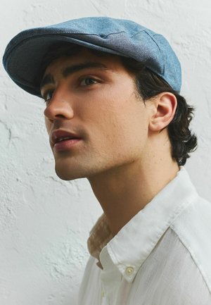 Junger Mann mit dunklem Haar, der eine blaue Schiebermütze und ein weißes Hemd mit Knöpfen trägt, und leicht nach oben blickt, vor einer strukturierten weißen Wand.