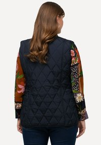 Marineblaue Steppweste mit tailliertem Design, ausgestattet mit einem hohen Kragen und seitlichen Nähten. Das darunter tragende Hemd mit Blumennärmeln zeigt lebendige Muster in Herbstfarben.