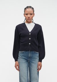 Young person with braided hair wearing a black buttoned cardigan over a white top and blue jeans, standing against a plain white background.