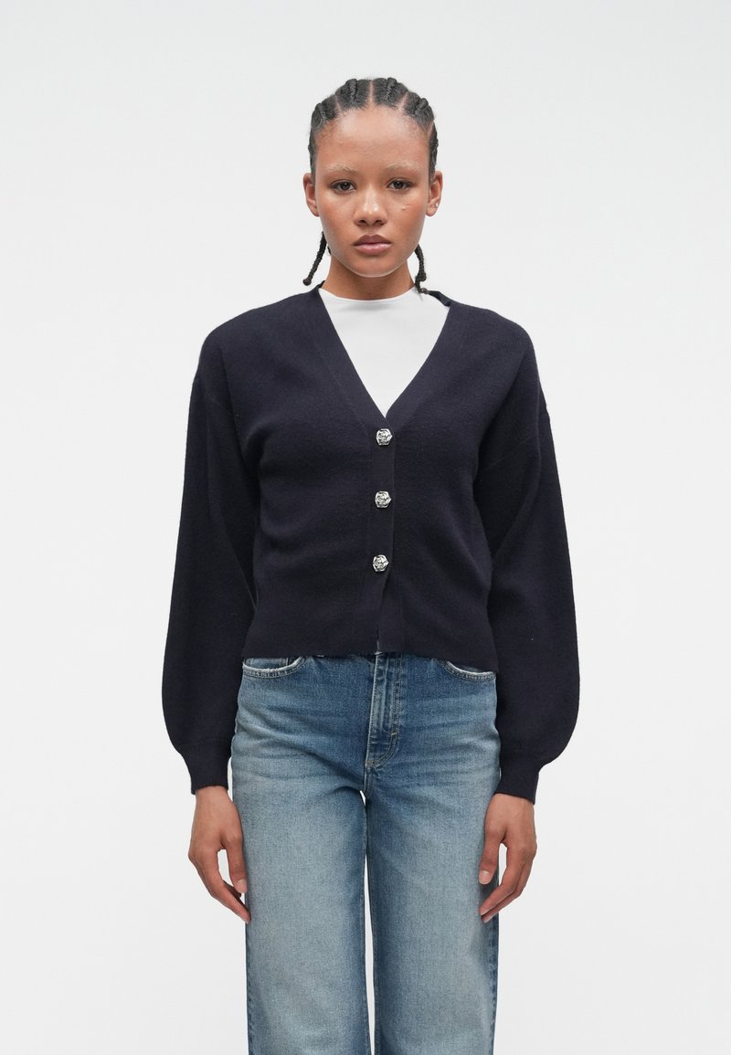 Young person with braided hair wearing a black buttoned cardigan over a white top and blue jeans, standing against a plain white background.