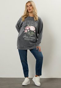 Sudadera gris de gran tamaño con gráfico floral y texto, combinada con jeans azules y zapatillas blancas, que cuenta con un corte relajado y puños acanalados.
