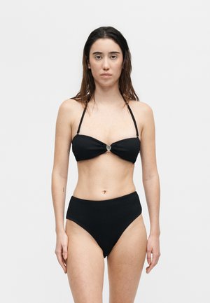 Femme aux longs cheveux foncés portant un bikini deux pièces noir avec un haut bandeau et un bas taille haute, debout devant un fond clair uni.