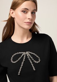 Tricou negru cu un mare design de fundă strălucitoare din cristale pe piept, completat cu detalii intricate și o textură moale a țesăturii.