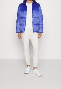 Blå vadderad jacka med glansigt ytskikt, dragkedja och tvåfärgad design. Matchad med cremefärgade mjukisbyxor och vita sportskor.