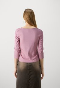 Pitkähihainen, pyöreäkauluksinen paita pehmeässä vaaleanpunaisessa kankaassa. Hivenen vartalonmyötäinen, jossa on kolme neljännestä pituista hihaa, yhdistettynä polvipituiseen ruskeaan hameeseen.