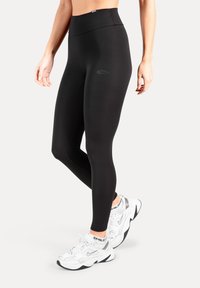 Zwarte leggings met hoge taille, gemaakt van een gladde, rekbare stof en voorzien van een subtiel logo op de dij, gecombineerd met witte sportschoenen.