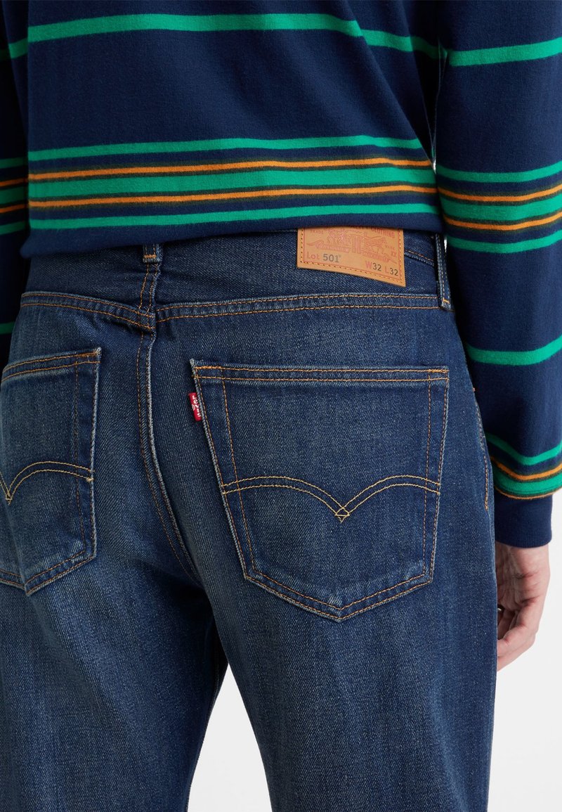 Dunkelblaue Jeans aus Denim mit klassischer Form, ausgestattet mit Gesäßtaschen mit orangefarbener Stickerei und einem braunen Lederpatch, das mit "Lot 501 W32 L32" beschriftet ist.