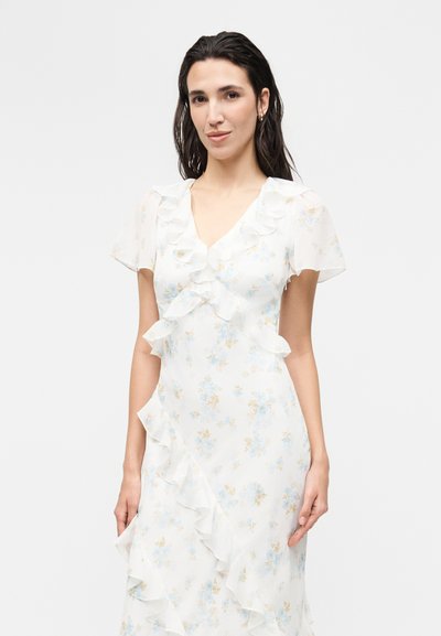 Γυναίκα με σκούρα μαλλιά που φοράει λευκό floral φόρεμα με βολάν στο λαιμό V και φαρδιά μανίκια, στέκεται μπροστά σε απλό φωτεινό φόντο.