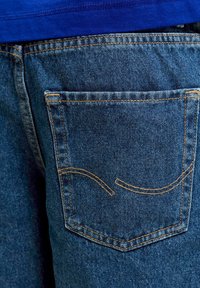 Πίσω τσέπη από μπλε τζιν με κίτρινη ραφή και ένα μπλε πουκάμισο μερικώς ορατό πάνω από τη μέση.