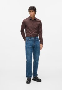 Camicia marrone con bottoni, jeans in denim blu, mocassini neri. Tessuto morbido, vestibilità classica, design minimalista con dettaglio logo discreto sulla camicia.