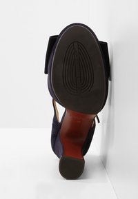 Une seule chaussure à talon haut bleu marine reposant sur une surface blanche montrant la semelle noire avec le nom de la marque et la pointe talon texturée.