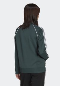 Dark green zip-up jacket with white accents, ribbed cuffs and hem, and diagonal white stripes on the shoulders. Soft, smooth texture.