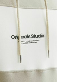 Beige og hvid hættetrøje med logoet "Originals Studio" i sort. Lavet af blødt stof med snore og sølvmetal spidser.