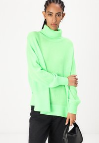 Neon grønn oversized genser med høy hals og ribbede mansjetter, sammen med svarte bukser og en liten svart håndveske.