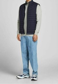 Tmavě modrá prošívaná vesta přes světle šedou mikinu na zip, v kombinaci s modrými džíny s rovnými nohavicemi a černo-bílými sportovními botami.