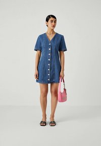 Mulher vestindo um vestido azul de ganga de manga curta com botões à frente, segurando uma mala rosa e usando sandálias pretas de slide sobre um fundo simples.