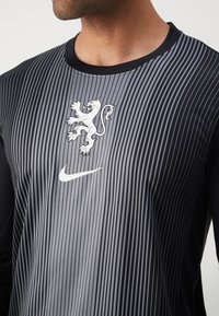 Zwart en grijs sportshirt met lange mouwen met verticale strepen, een witte leeuwenlogo en een witte Nike swoosh op de voorkant.