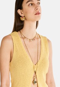 Chaleco de punto amarillo con un escote en V profundo, combinado con cadenas de oro en capas que tienen colgantes en forma de corazón y charms, y grandes aretes de oro.