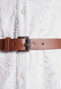 Ceinture en cuir marron fermée par une boucle argentée autour de la taille sur une chemise blanche à boutons avec broderie à motif de feuilles.