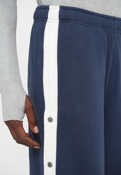 Close-up van marineblauwe sportbroek met witte zijkant en drukknoopjes, gedragen met een grijze longsleeve shirt met duimgaten.