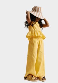 Žlutý lněný dvoudílný outfit obsahující peplum top a široké kalhoty, doplněný růžovo-bílým pruhovaným kloboukem s kšiltem.
