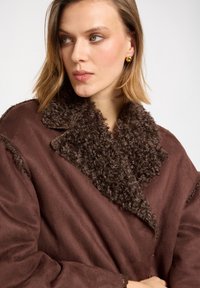Manteau marron avec un col en fourrure brun foncé, texture lisse, design surdimensionné et manches larges. Les détails incluent des bords effilochés.