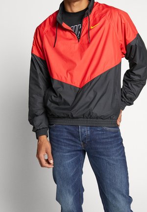 Mand iført en rød og sort vindjakke med lynlås over en sort Nike-shirt og blå jeans, stående med den ene hånd i lommen.