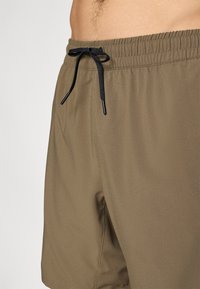 Καφέ μαγιό με ελαστική μέση, μαύρο κορδόνι και λεία υφή. Ορατές πλαϊνές τσέπες στο εξωτερικό ύφασμα.