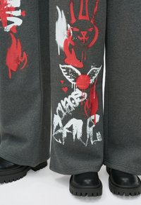 Graue Sweatpants mit weitem Bein, verziert mit roten und weißen grafischen Designs, darunter ein Hase, Flügel und Textelemente. Texturiertes Gewebe mit lockerem Sitz.